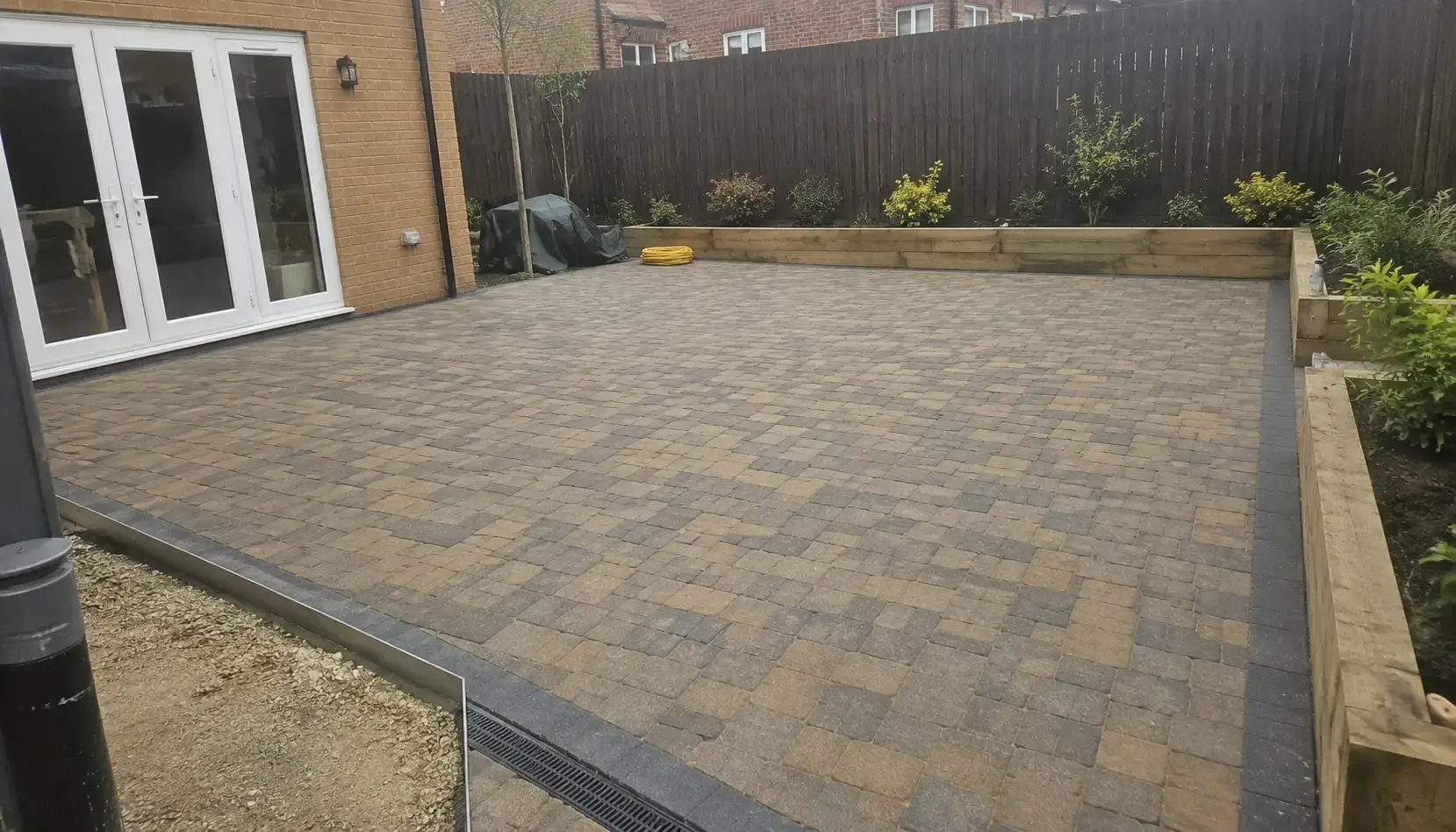 Newly installed block paved patio in Ely, Cambridgeshire with charcoal border edging, raised timber planters and modern brick garden setting.