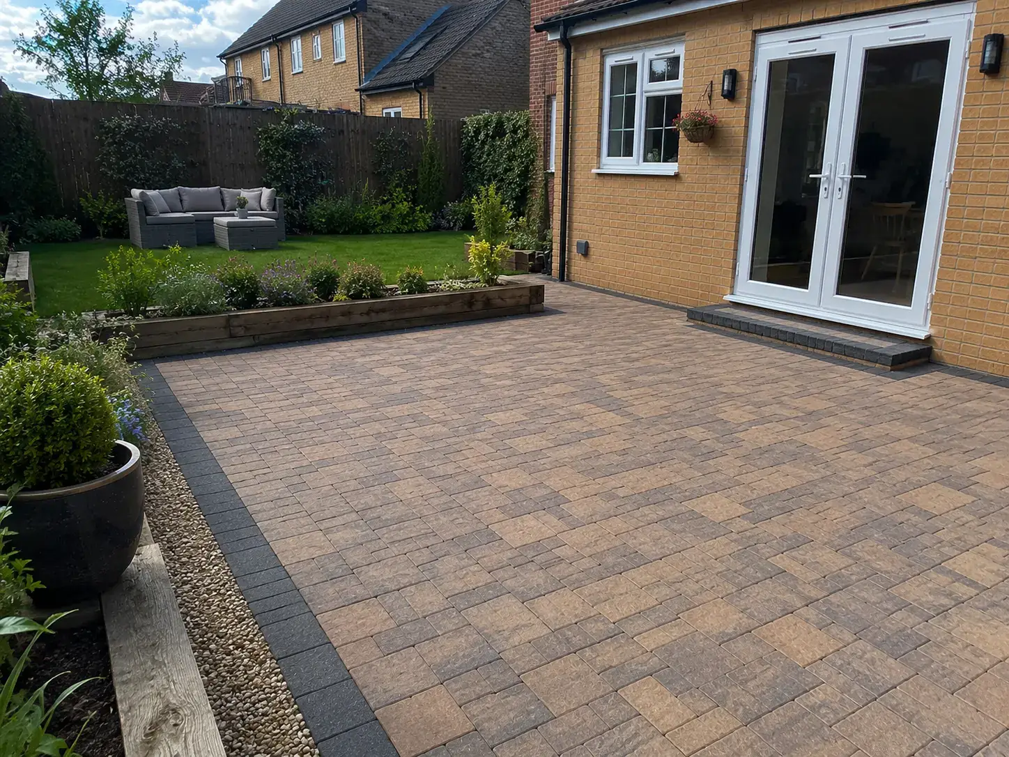 Completed block paved patio in Ely, Cambridgeshire featuring a charcoal border, landscaped garden beds and a modern brick home.