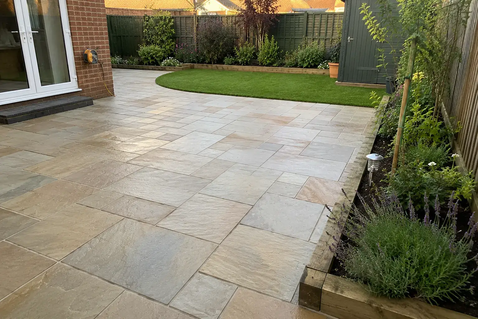 Large Indian sandstone patio with landscaped garden, curved lawn, raised flower beds and modern brick home in Ely, Cambridgeshire.