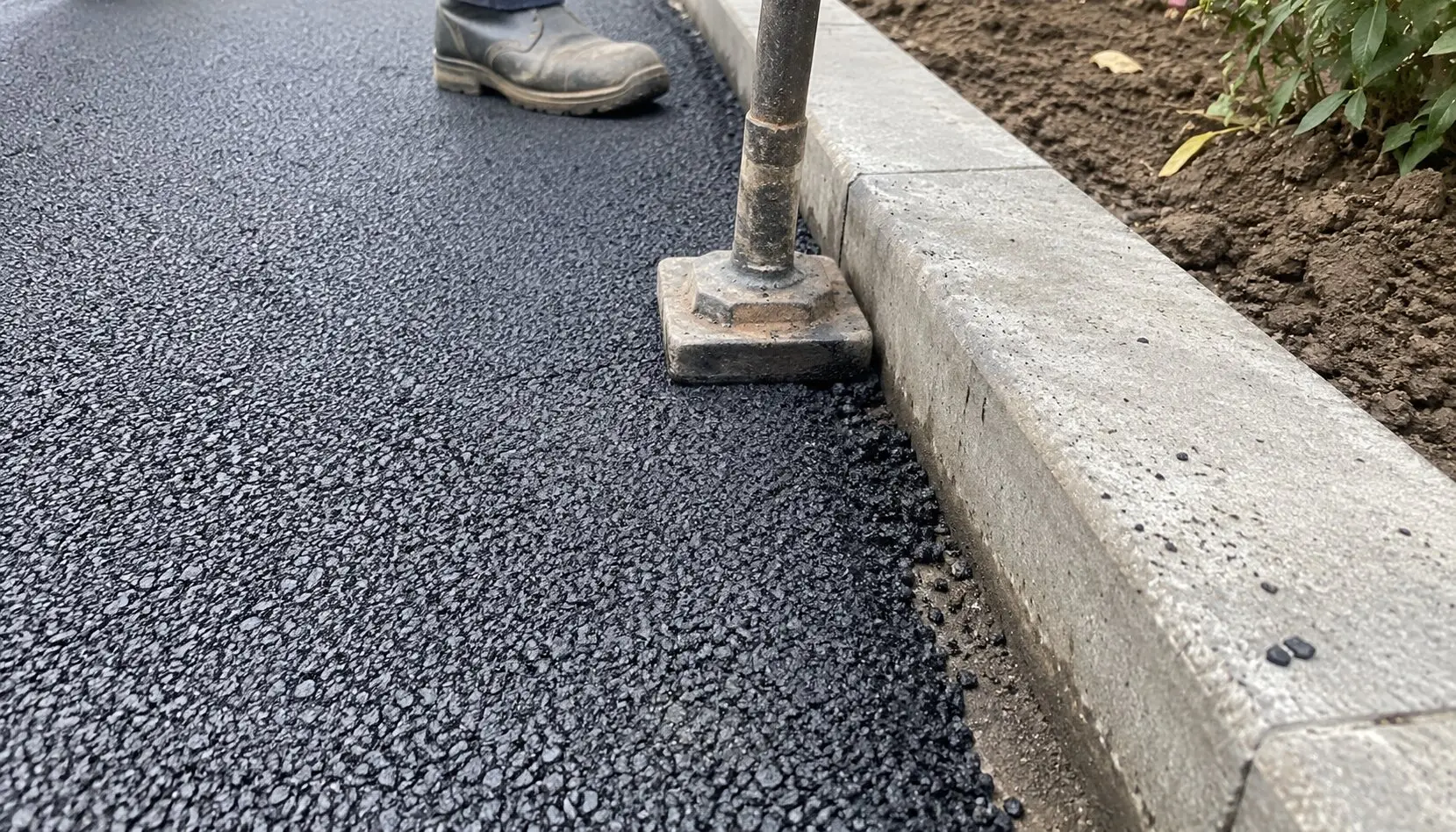Tarmac driveway installation in Ely, Cambridgeshire with fresh tarmac compacted against a concrete kerb stone using a steel tamping tool.